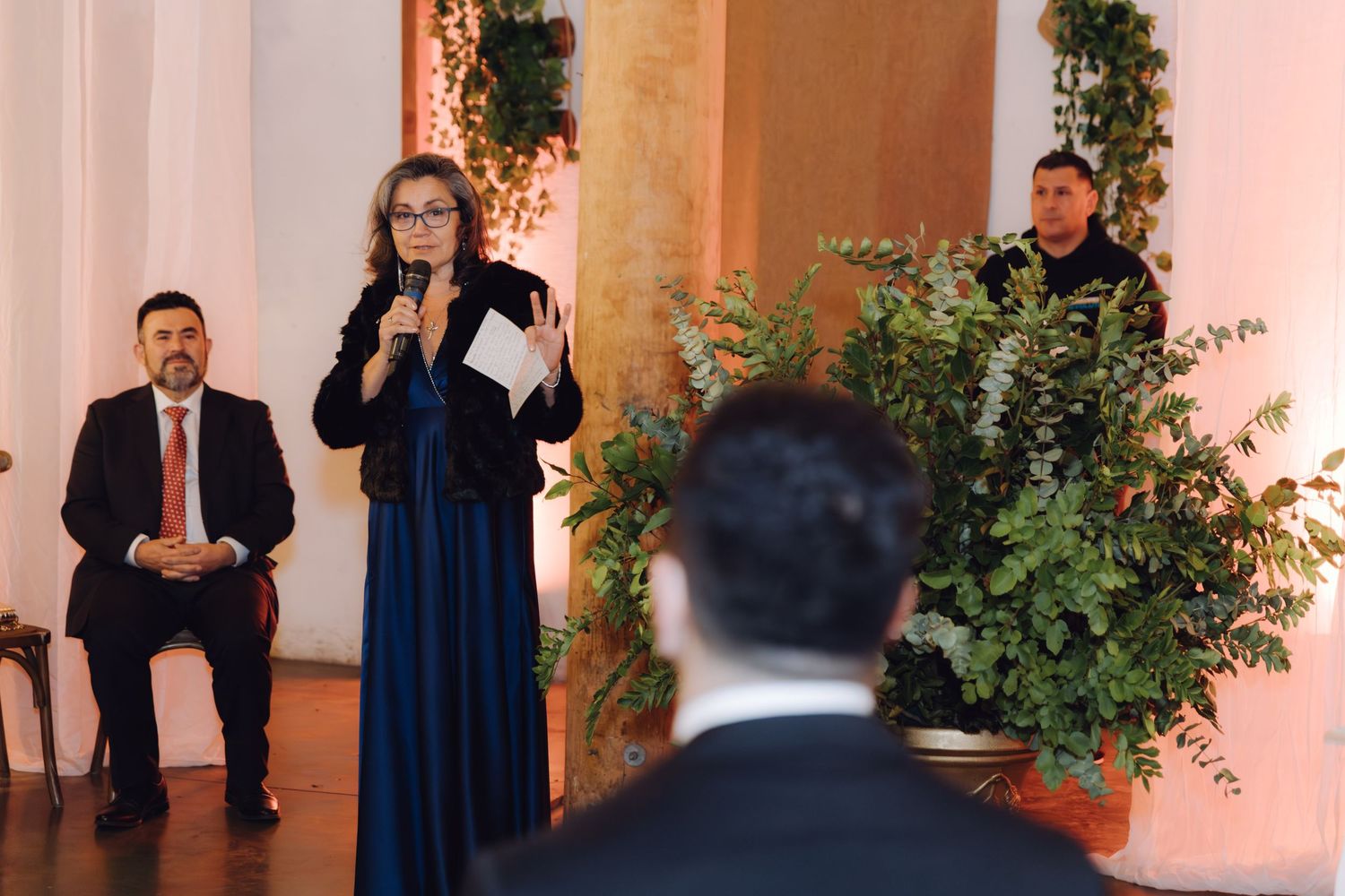 Woman in blue gown speaks at formal wedding ceremony while guests and officiant listen attentively indoors.