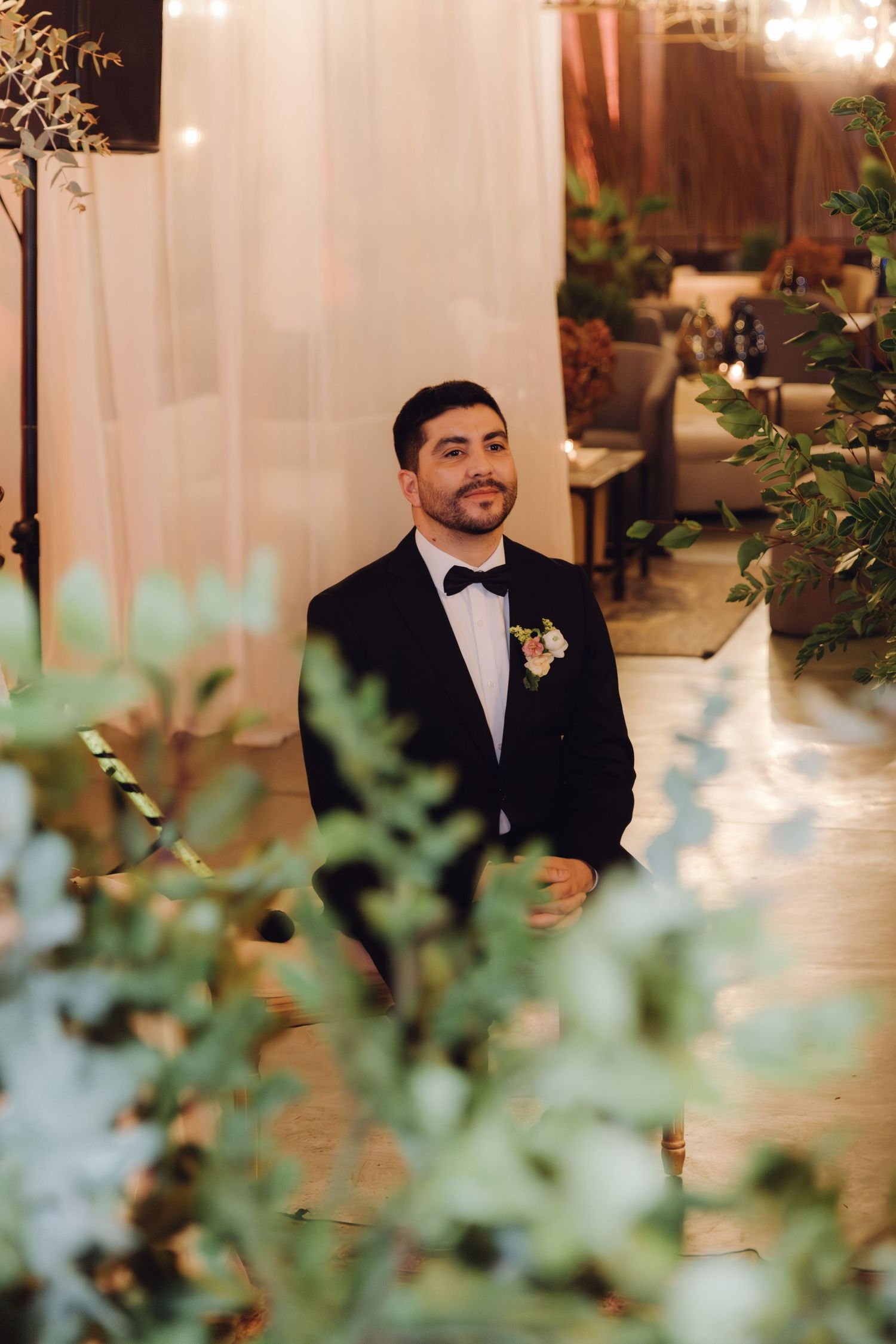 Handsome groom in black tuxedo with boutonniere stands at wedding altar surrounded by lush greenery and draping.