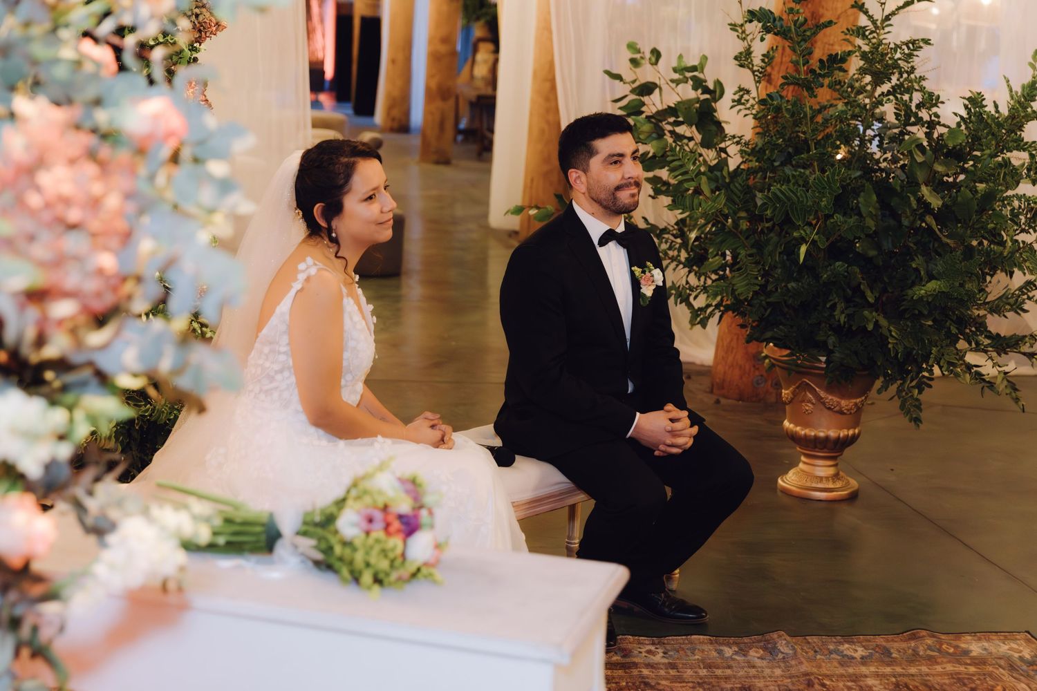 Bride and groom sit together at wedding ceremony altar with colorful floral bouquet and decorations nearby.