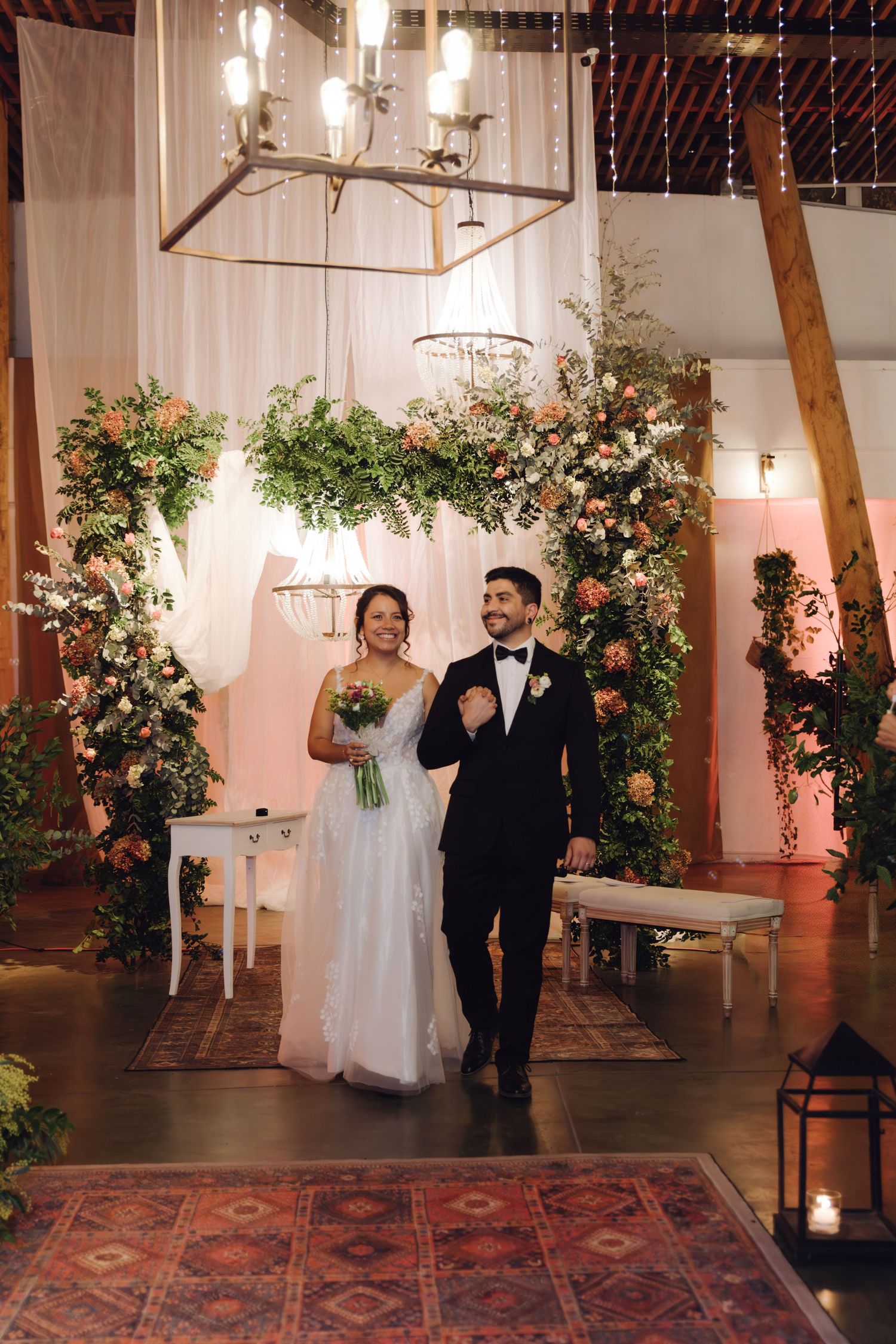 Joyful newlyweds walk down aisle after ceremony under chandelier with lush floral arch and warm lighting.