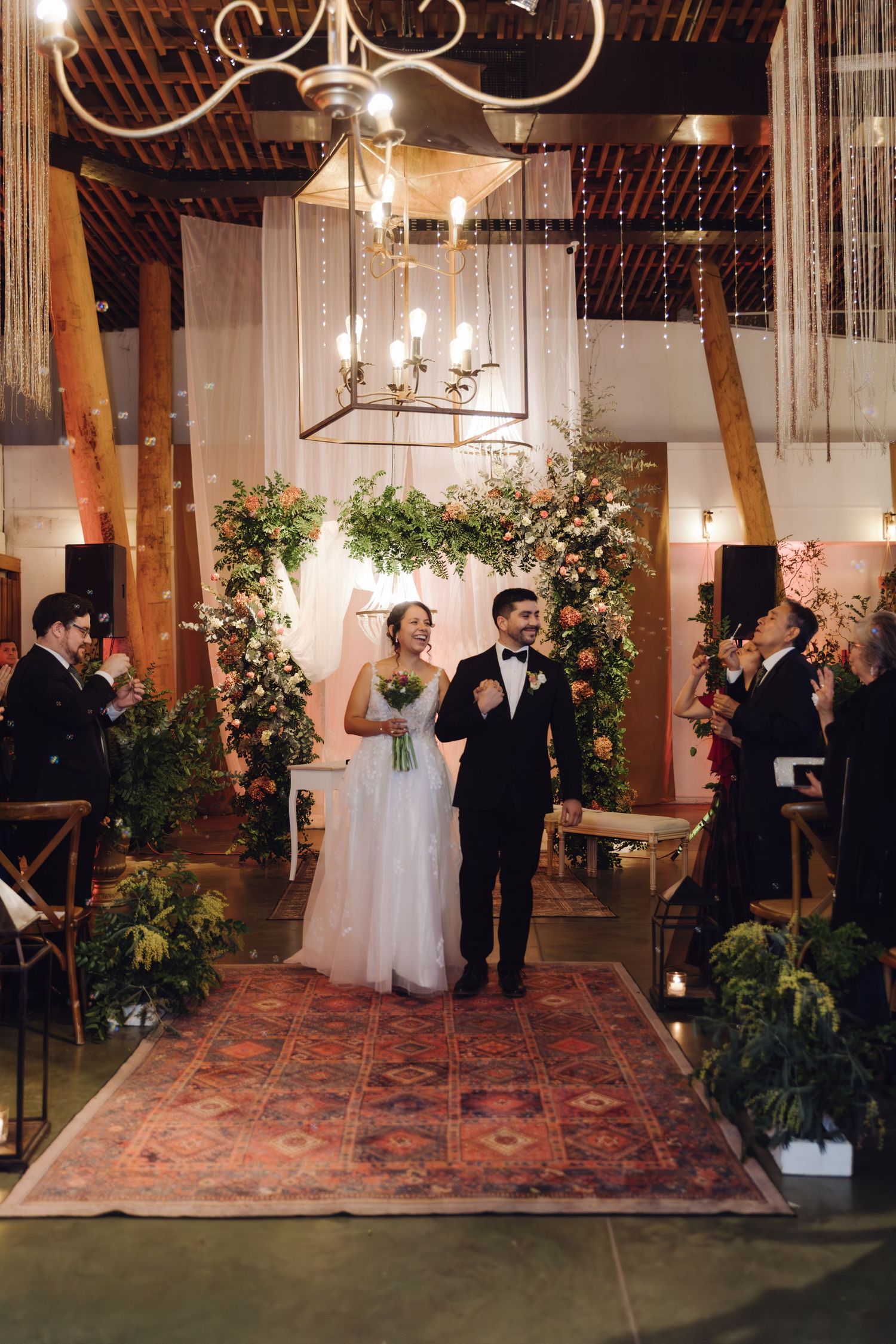 Newlyweds exit wedding ceremony aisle as guests celebrate with confetti under chandeliers and floral arch backdrop.
