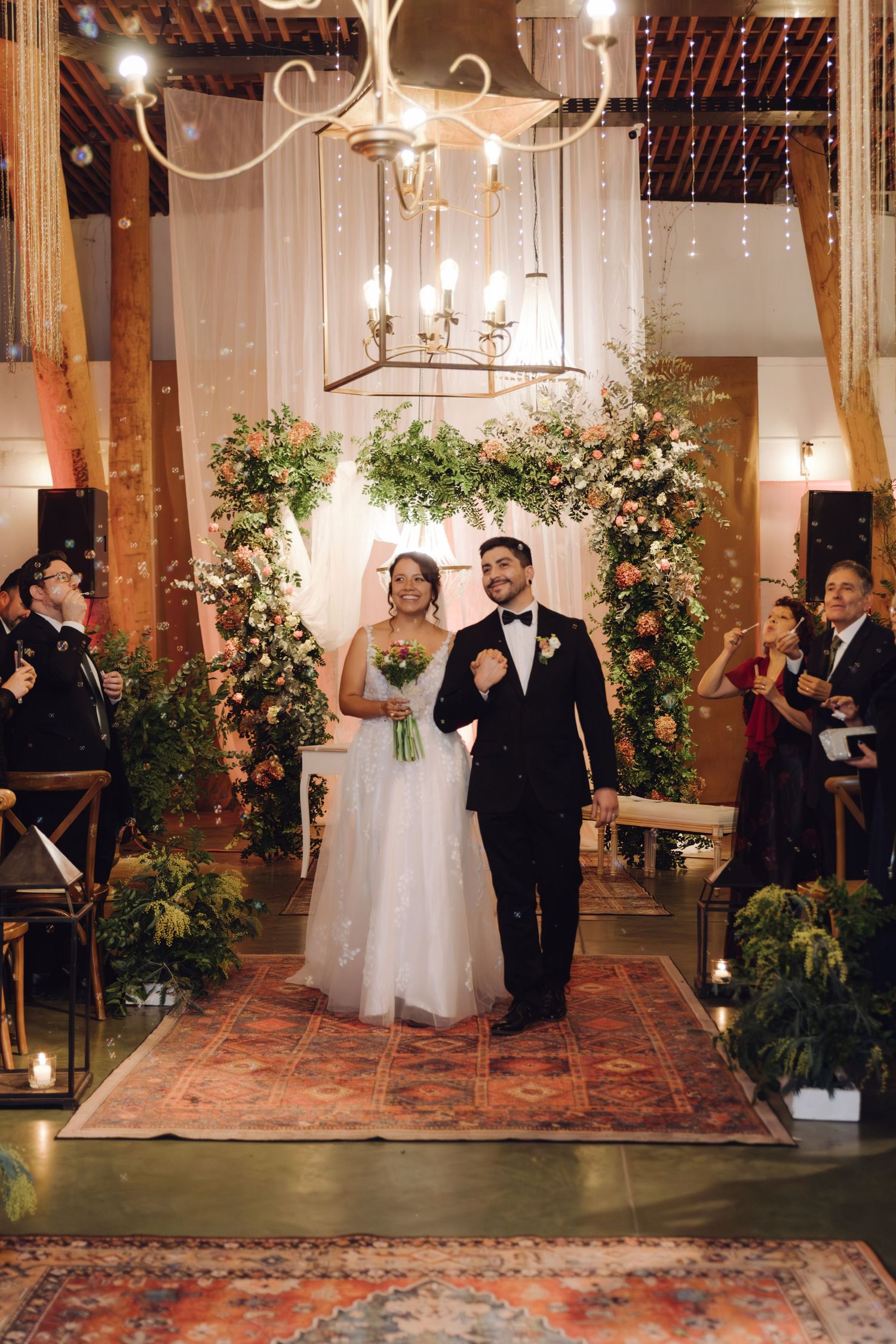 Happy newlyweds walk down aisle as guests cheer and toss confetti in warmly lit wedding venue.