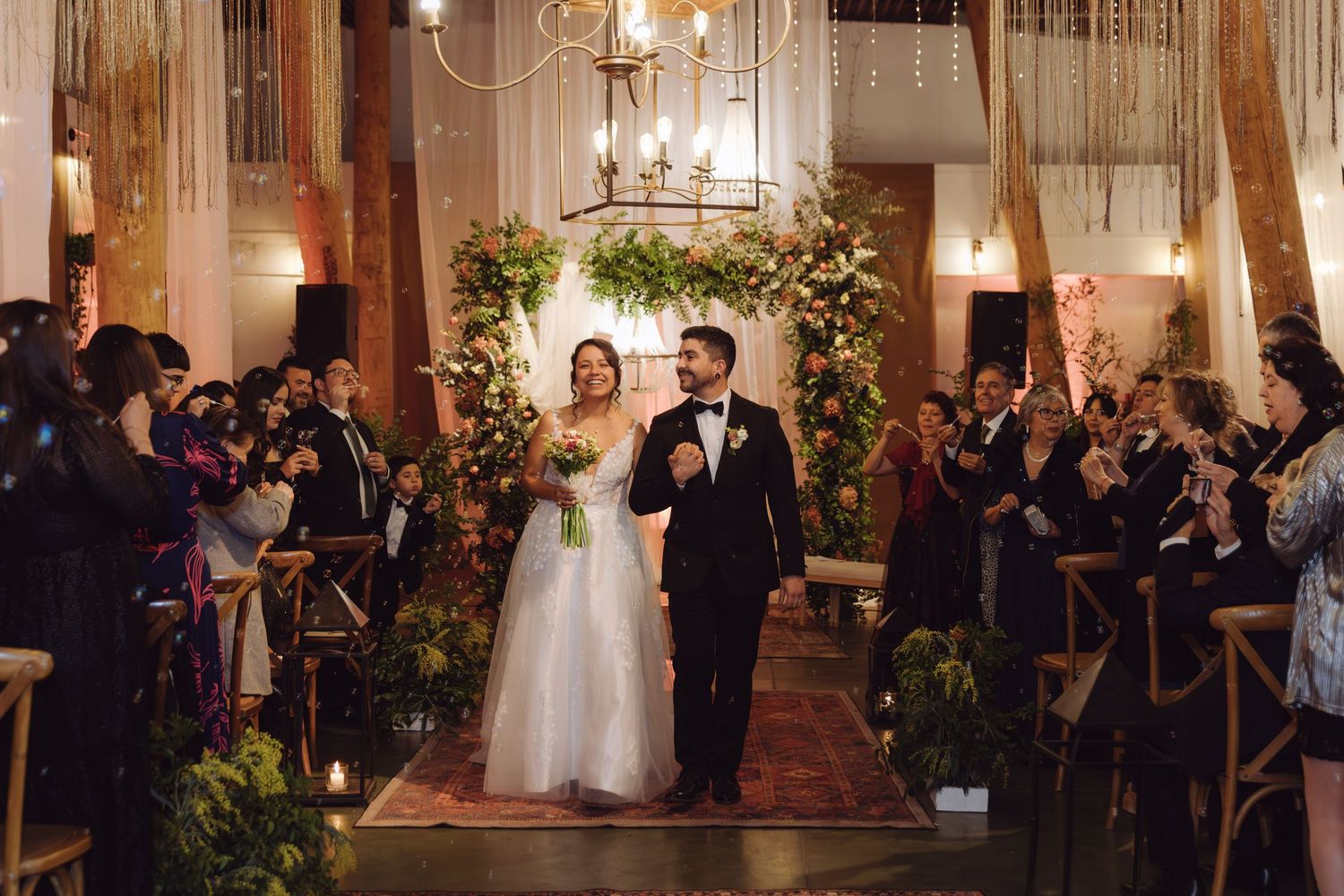 Smiling newlyweds walk down aisle together as guests celebrate in rustic warmly lit wedding ceremony venue.