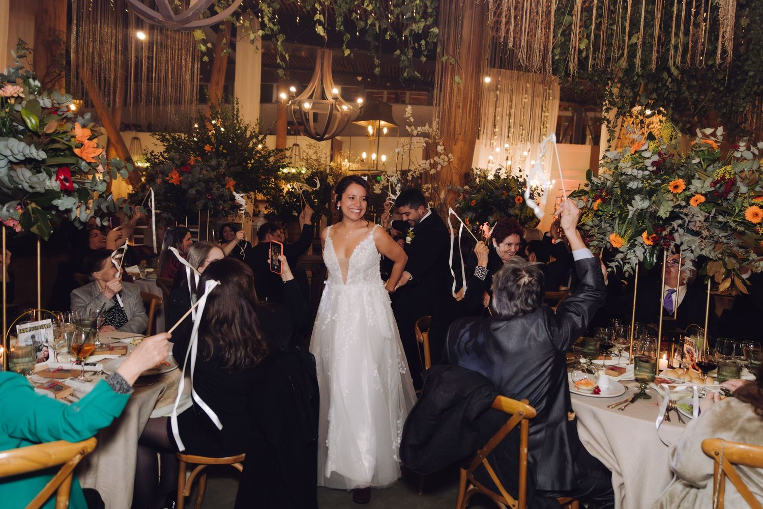 Bride walks through cheering guests at elegant wedding reception with lush floral arrangements and warm lighting.