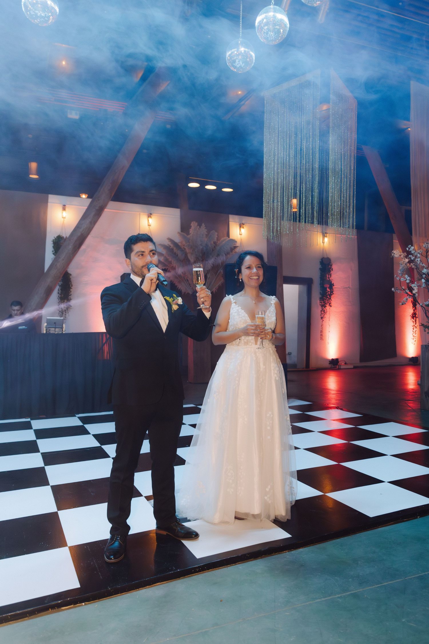 Newlyweds toast with champagne on checkered dance floor surrounded by atmospheric fog and soft lighting.