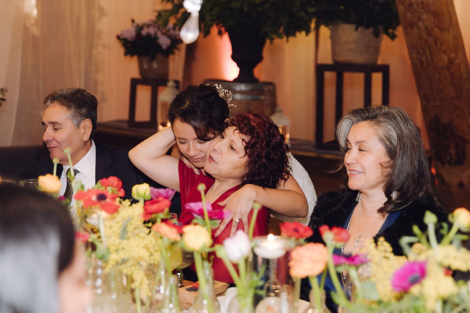 Wedding guests share emotional embrace at floral-decorated reception table with colorful blooms in foreground.