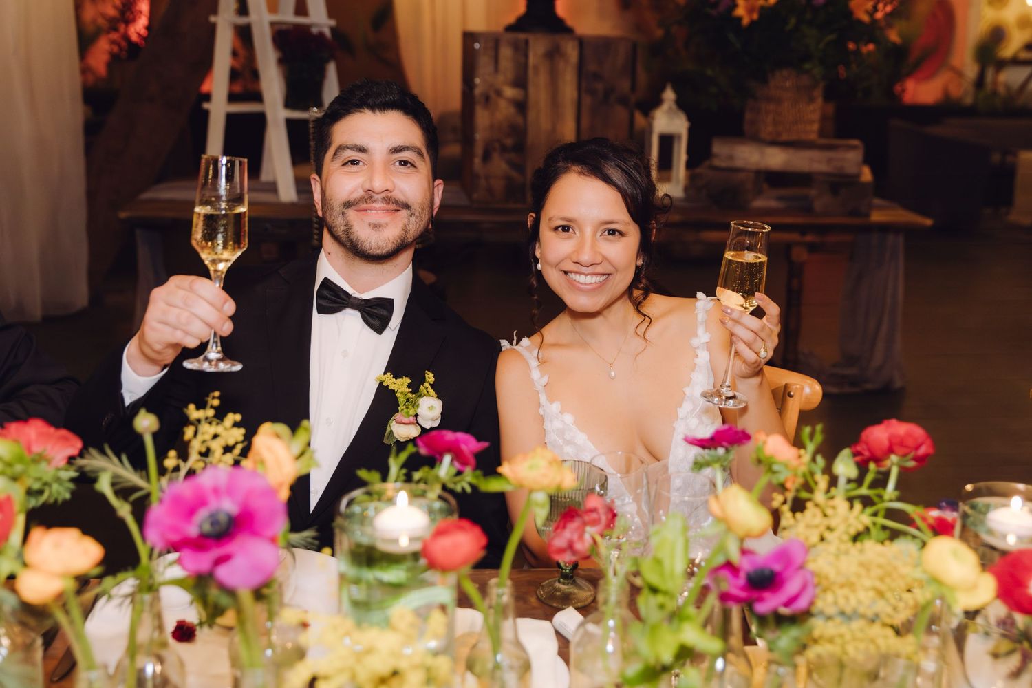 Smiling bride and groom raise champagne glasses at wedding reception table adorned with vibrant flowers.