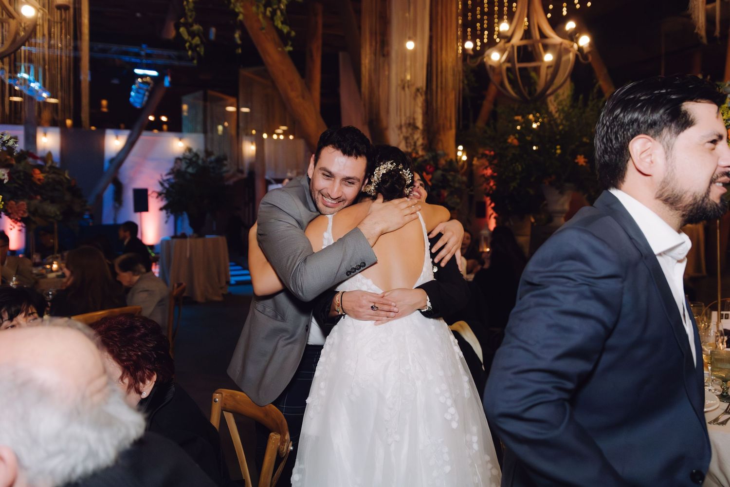 Groom embraces wedding guest warmly at beautifully decorated indoor reception with string lights overhead.