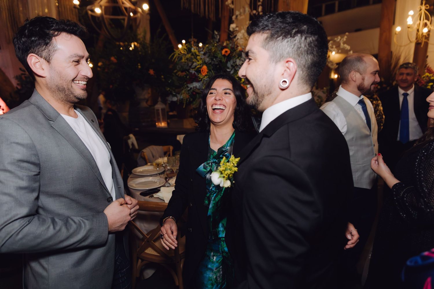 Three wedding guests laugh and socialize at elegant indoor reception with floral centerpieces in background.