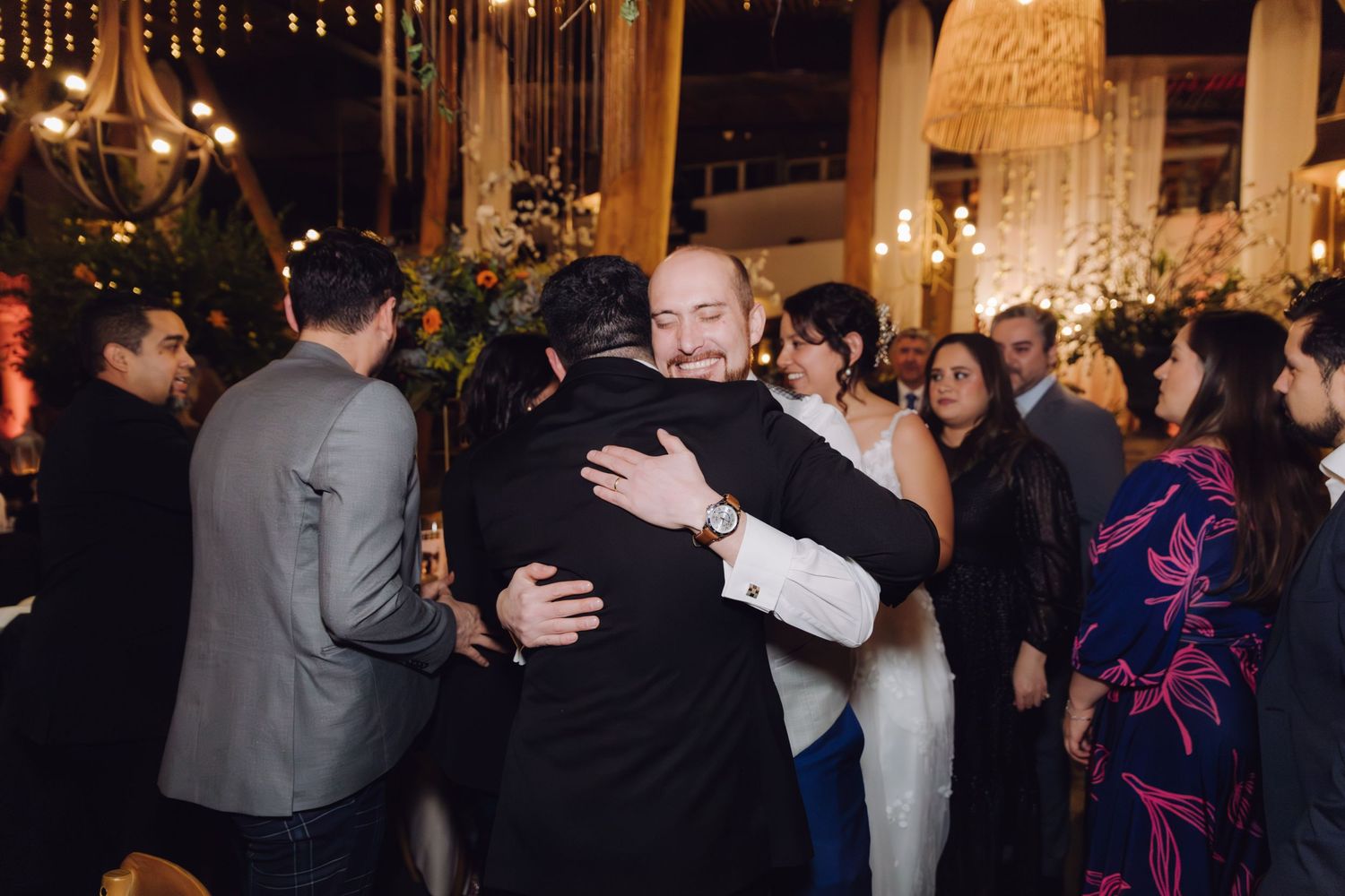 Wedding guests share heartfelt hugs at romantic indoor reception with glowing floral arrangements nearby.