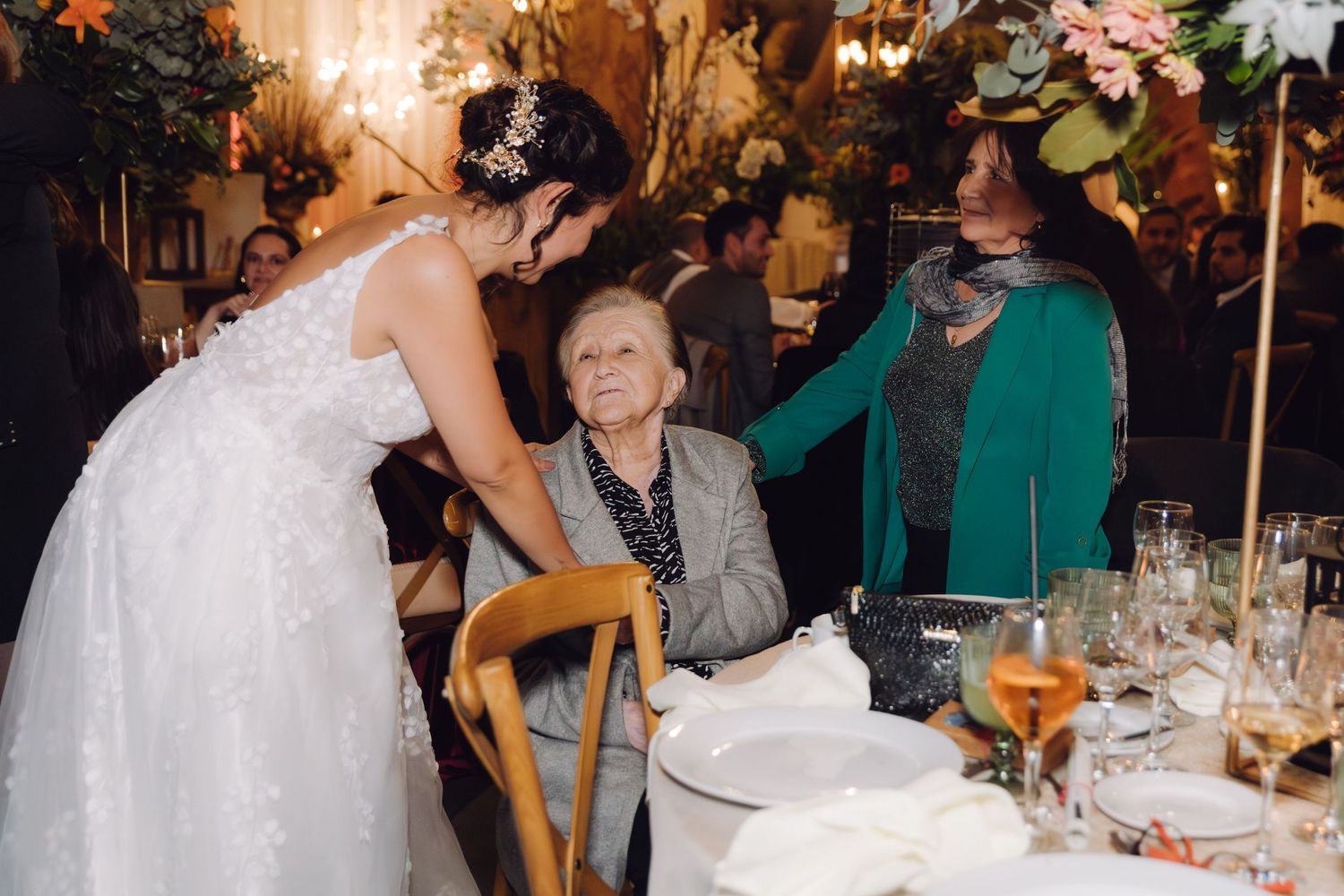 Bride leans down to greet elderly seated guest at elegantly decorated wedding reception table.