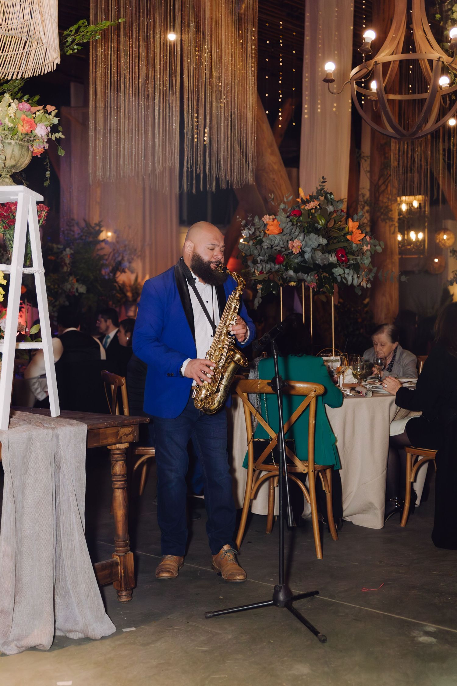 A saxophonist in a blue blazer performs at an elegant wedding reception with string lights and floral decorations.