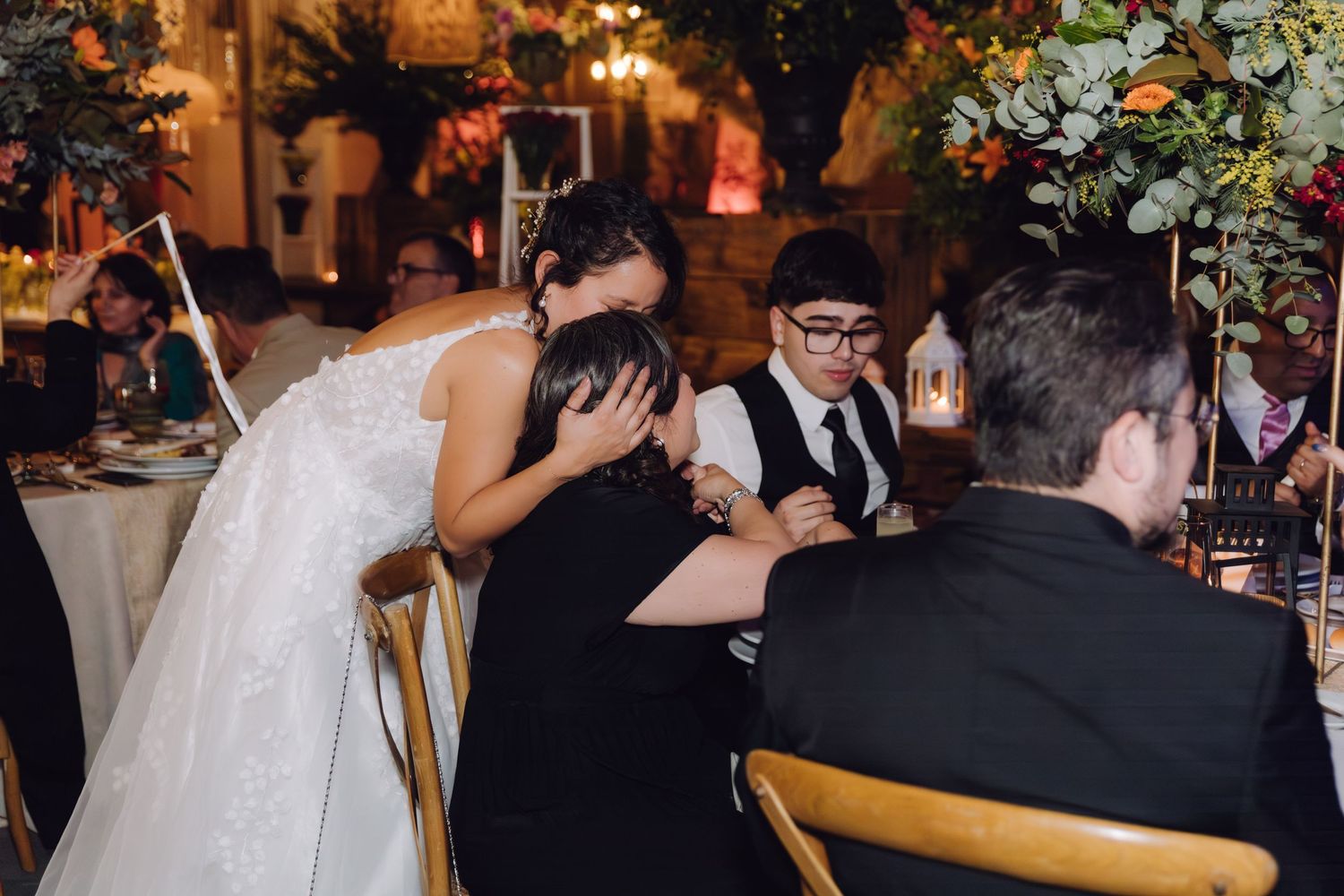A bride in a white lace gown embraces an emotional guest at a beautifully decorated wedding reception.