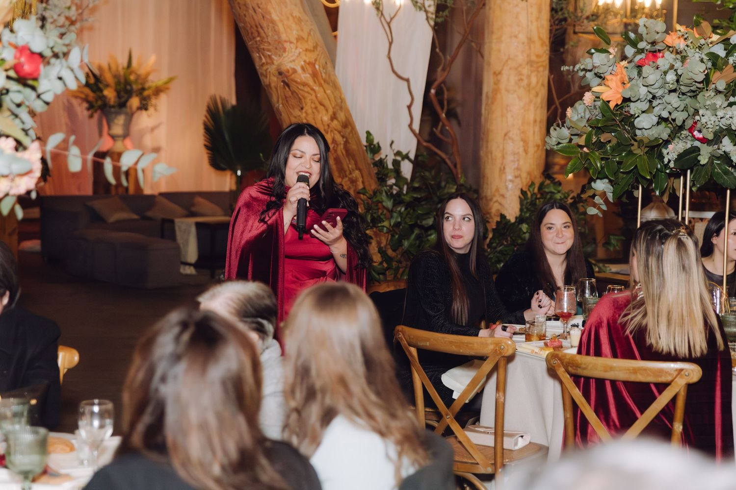 A woman in a red velvet dress speaks to guests at an indoor wedding reception with lush floral decor.