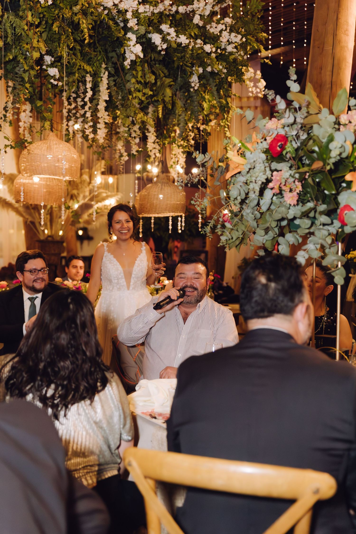 A bride smiles as a man speaks into a microphone at a wedding reception with greenery and string lights.