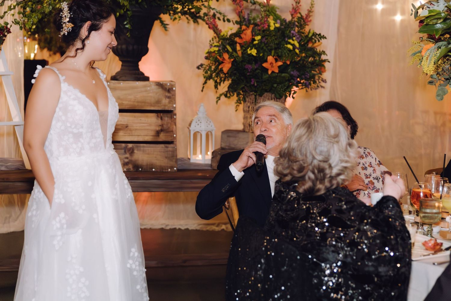 An older man kisses the hand of a bride in a lace gown at a warmly lit wedding reception.