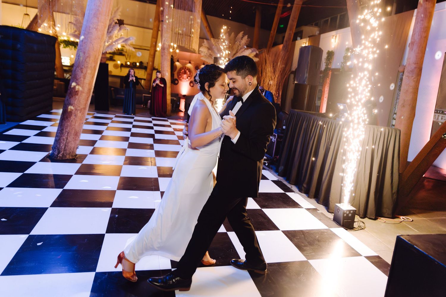 A couple shares a romantic first dance on a checkered floor with sparkling fireworks in the background.