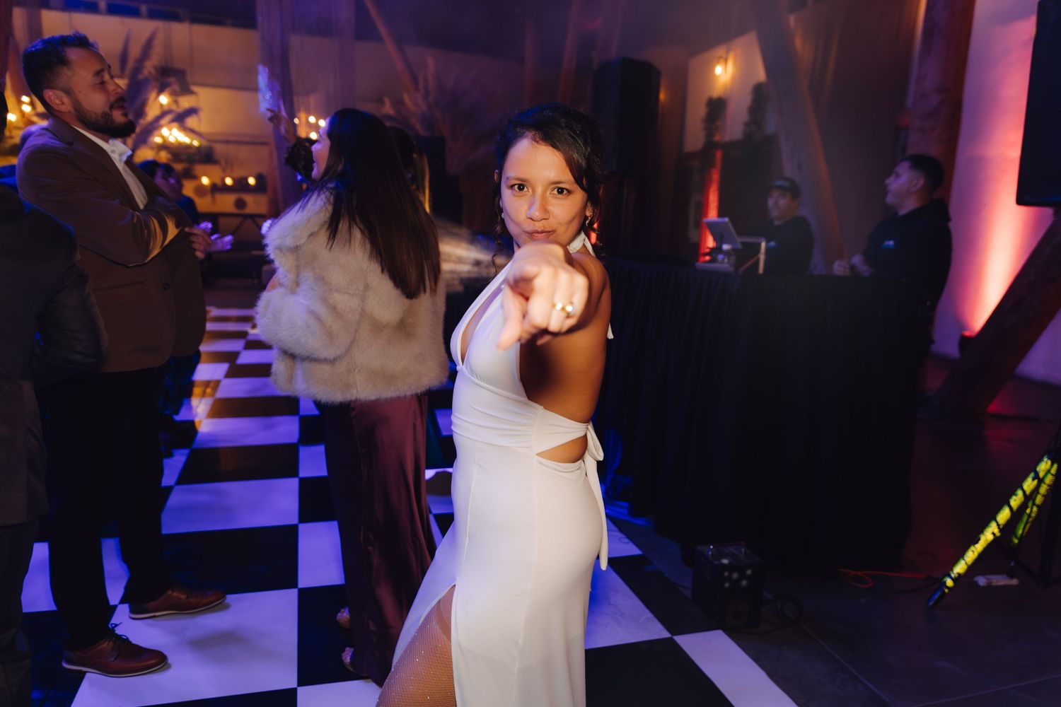 A bride in a white cutout gown poses playfully on a checkered dance floor at her wedding reception.