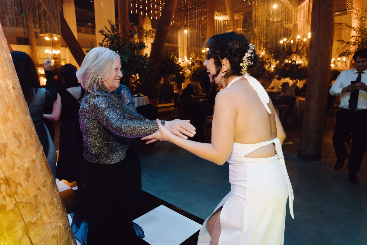 Bride in backless white gown holding hands with older woman at wedding reception.