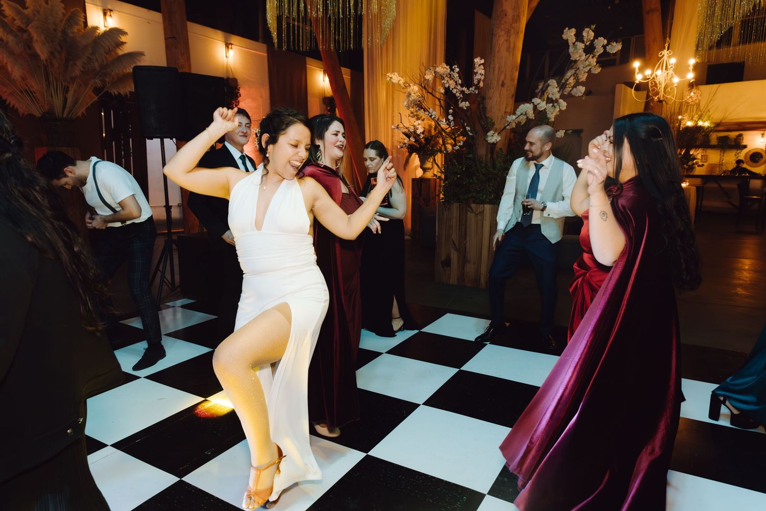Bride in white dress dancing energetically with bridesmaids in burgundy gowns on checkered floor.