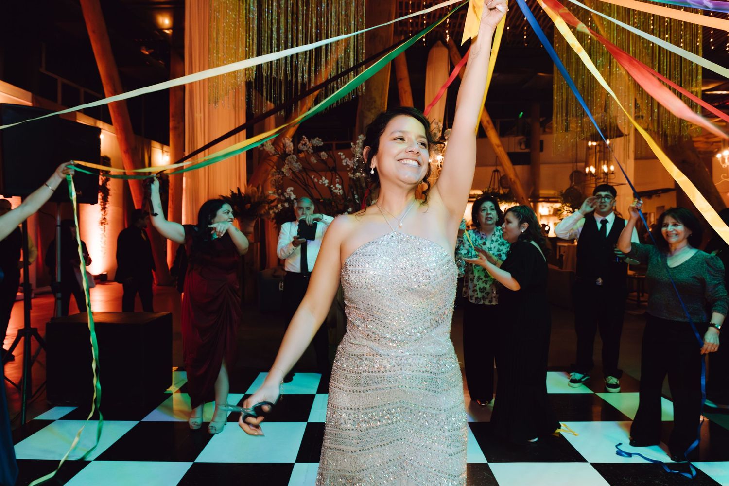 Bride in silver sequin gown dances joyfully on checkered dance floor with colorful streamers above.