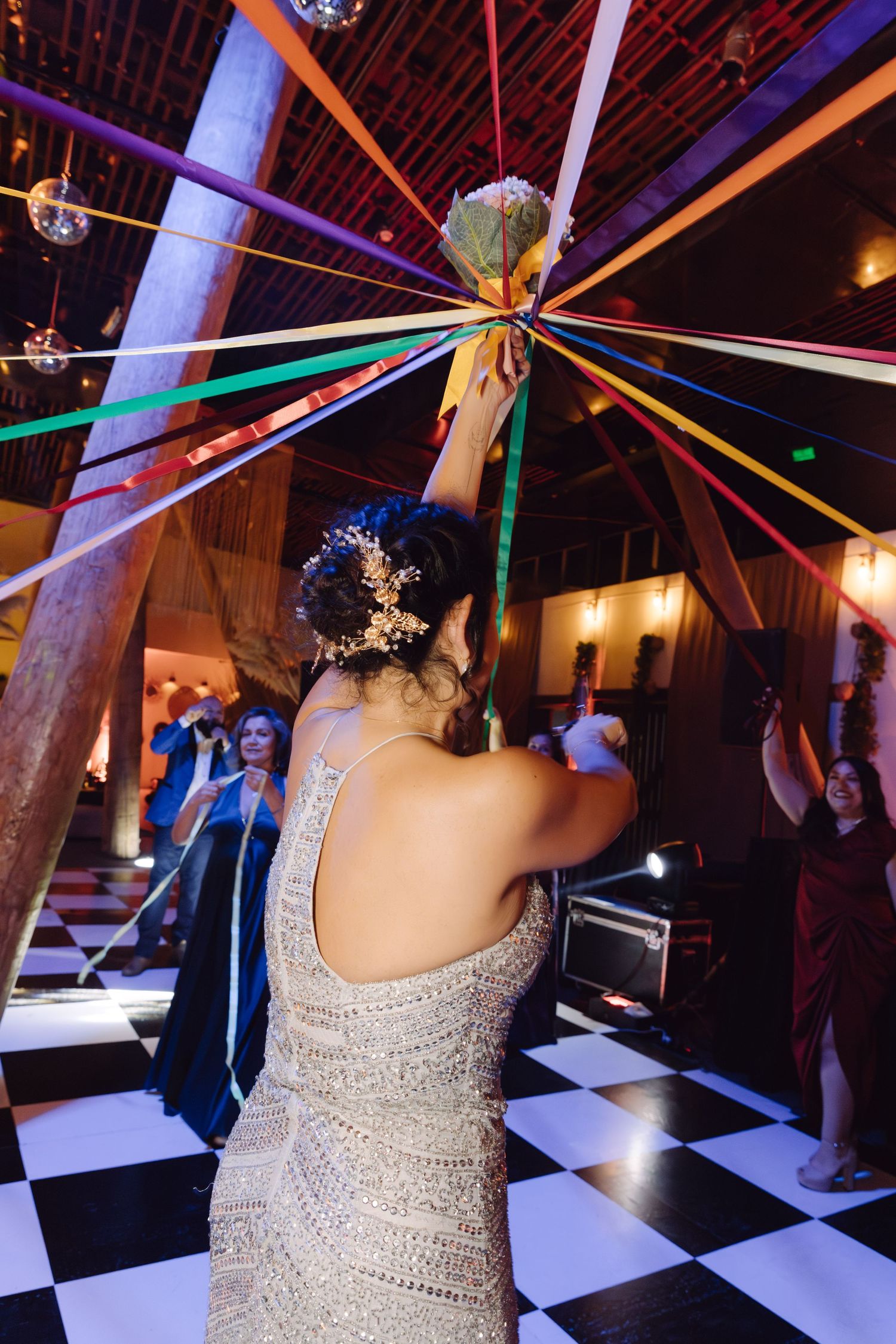 Bride in beaded silver gown raises bouquet amid vibrant colorful streamers on a checkered dance floor.