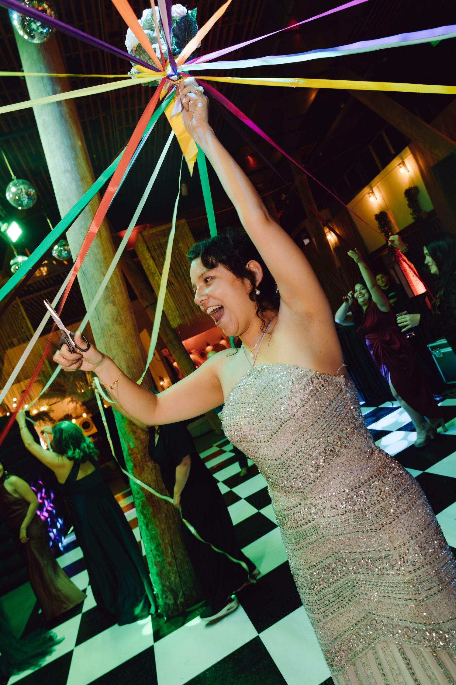 Woman in elegant silver dress laughing while holding colorful streamers at a lively wedding party.