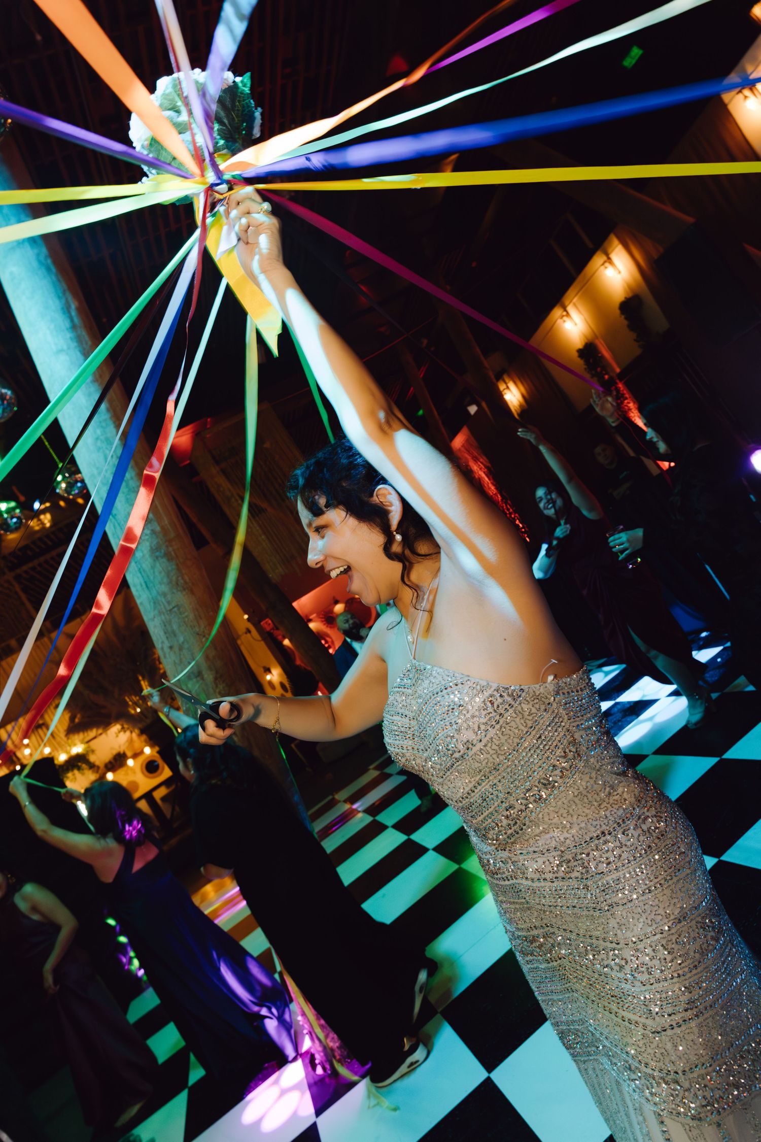 Bride in silver beaded gown dancing joyfully with colorful ribbons at a festive wedding reception.