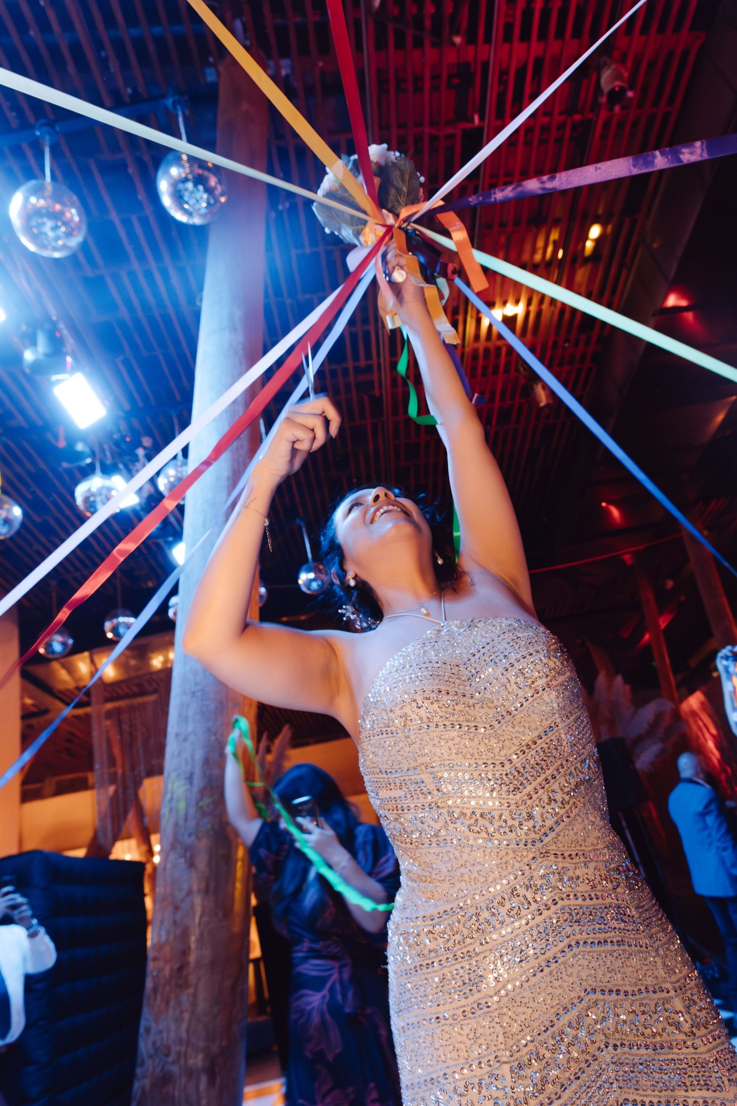Bride in sparkly gown reaching up to hold colorful ribbons under disco balls at wedding reception.