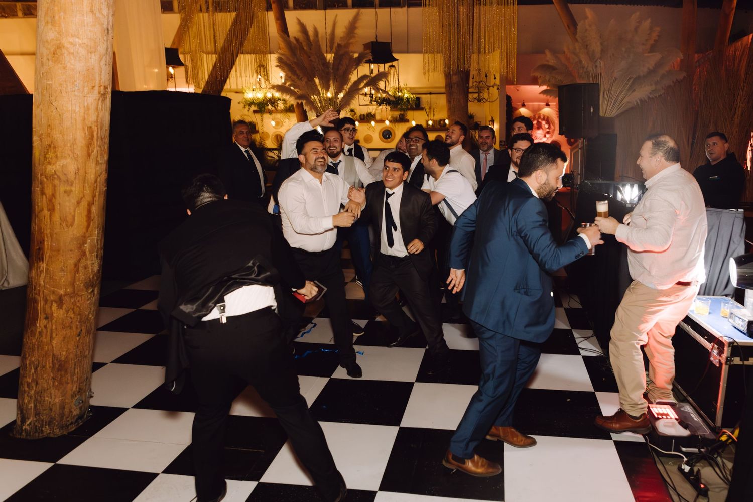 Energetic group of men in suits dancing on black and white checkered floor at wedding reception.