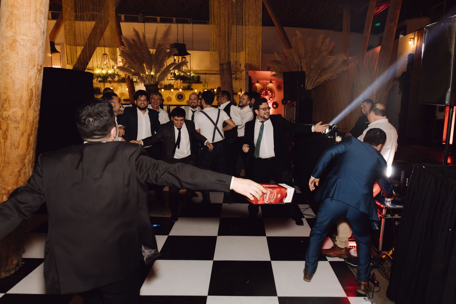 Groomsmen in black suits dancing energetically on checkered dance floor at a festive wedding party.