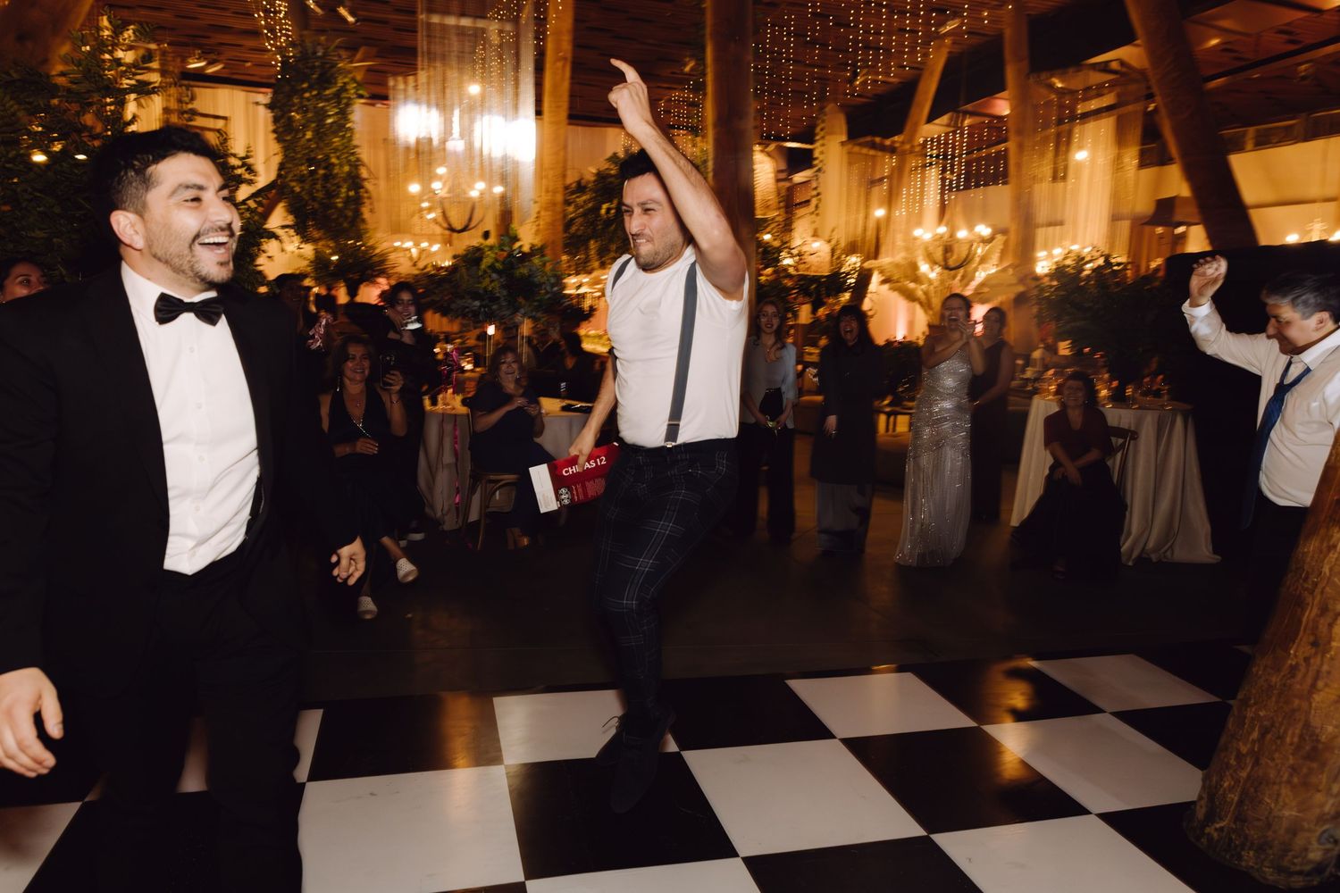 Cheerful man in suspenders high-kicking on black and white dance floor at elegant wedding reception.
