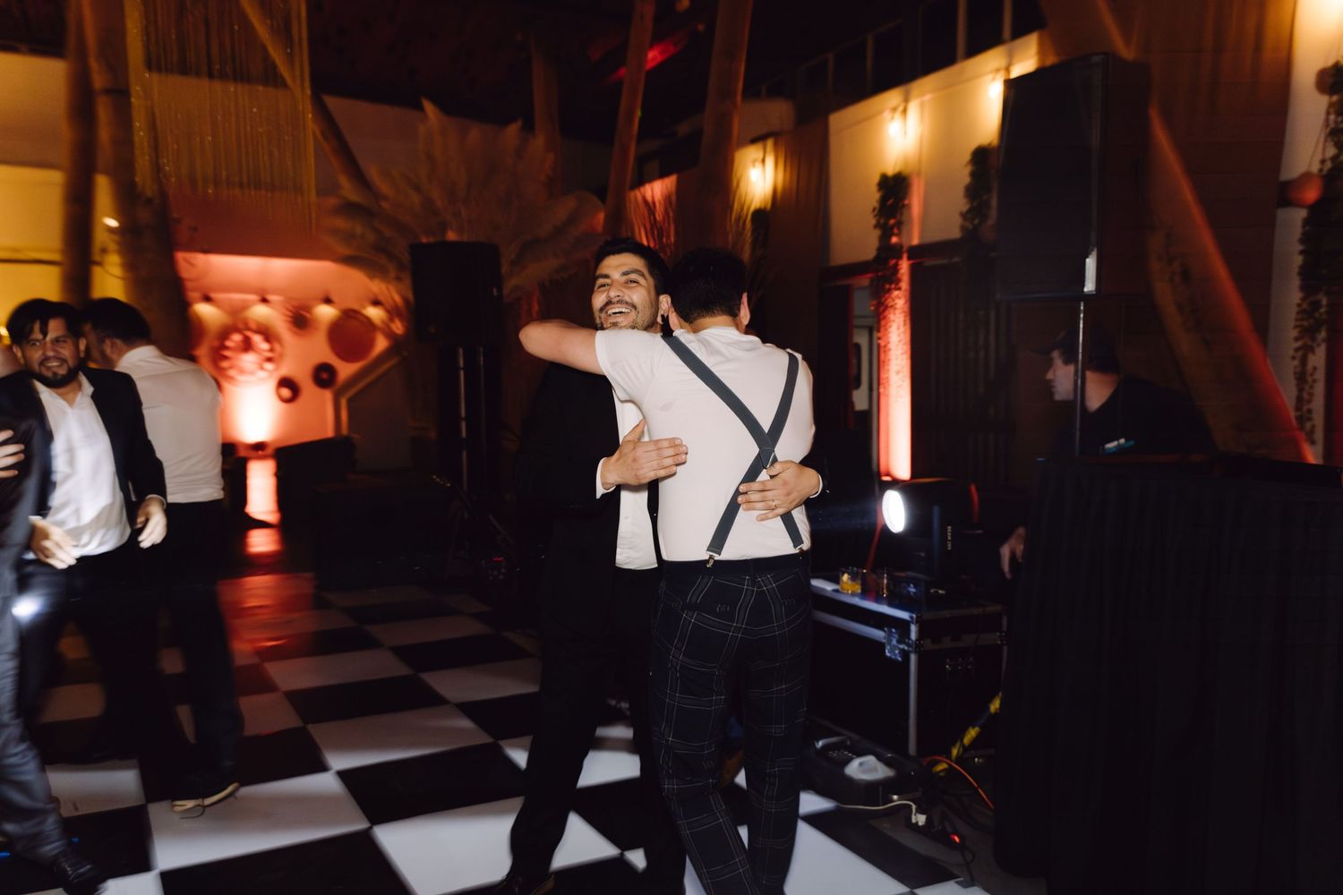 Two men in formal attire hugging and dancing on checkered floor at an outdoor wedding reception.