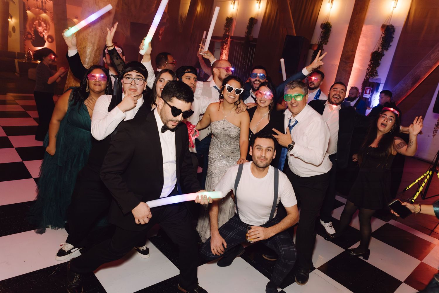 Large group of wedding guests posing with glow sticks and fun accessories on checkered dance floor.