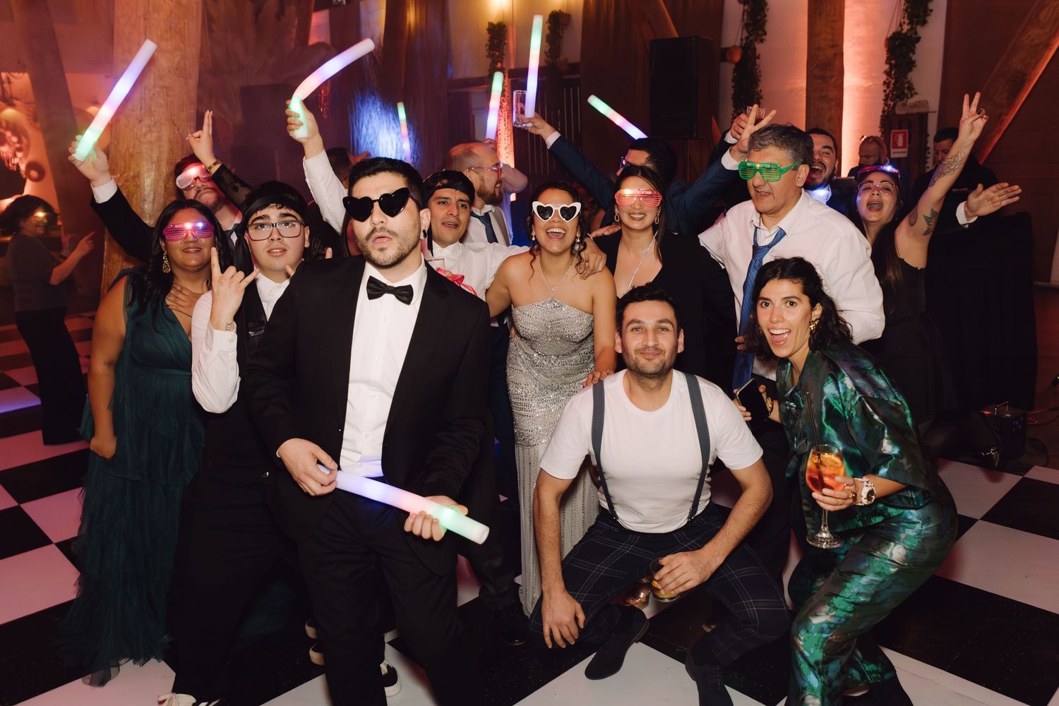 Wedding guests in formal wear posing with glow sticks and masquerade masks on checkered dance floor.