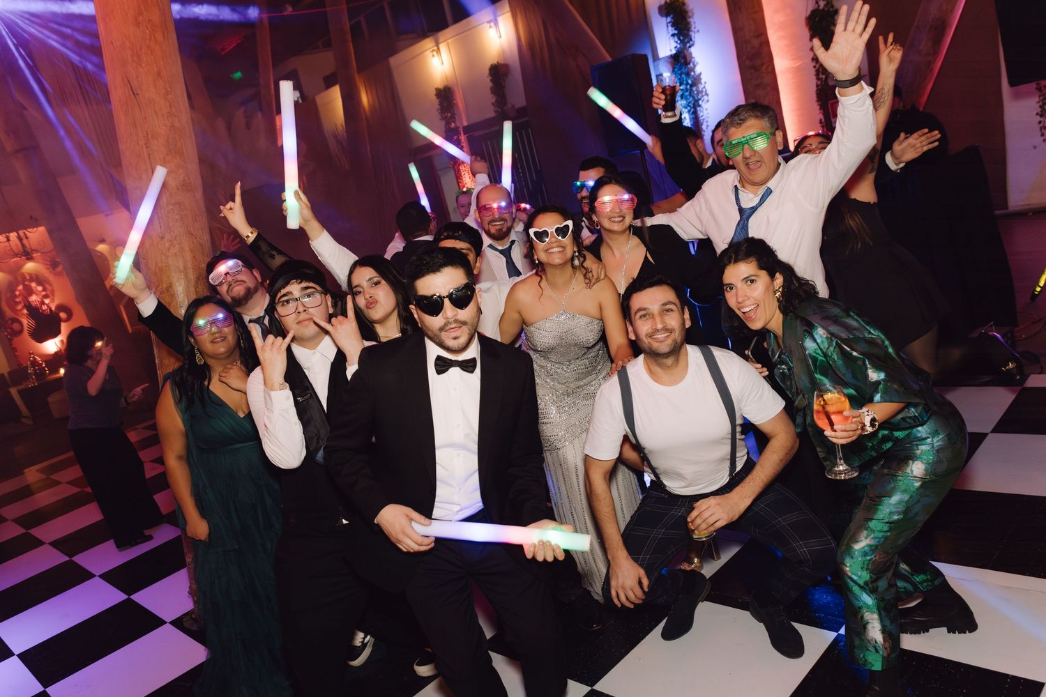 Smiling wedding party group wearing fun accessories and holding glow sticks at festive reception.
