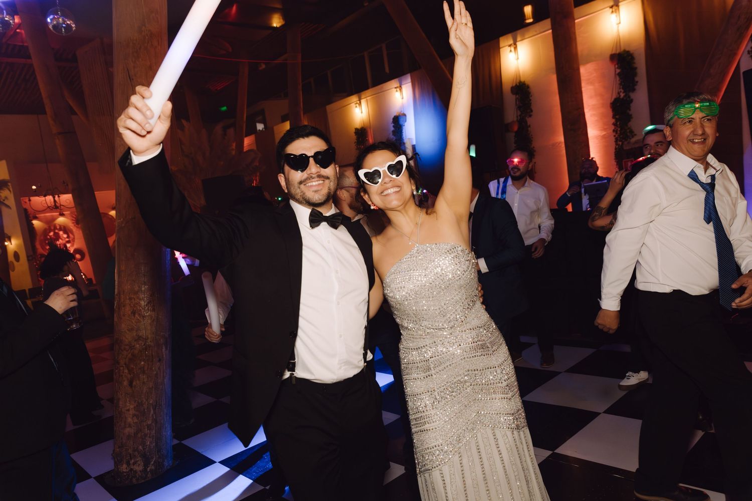 Bride and groom dancing joyfully at wedding reception wearing sunglasses on black and white checkered dance floor.
