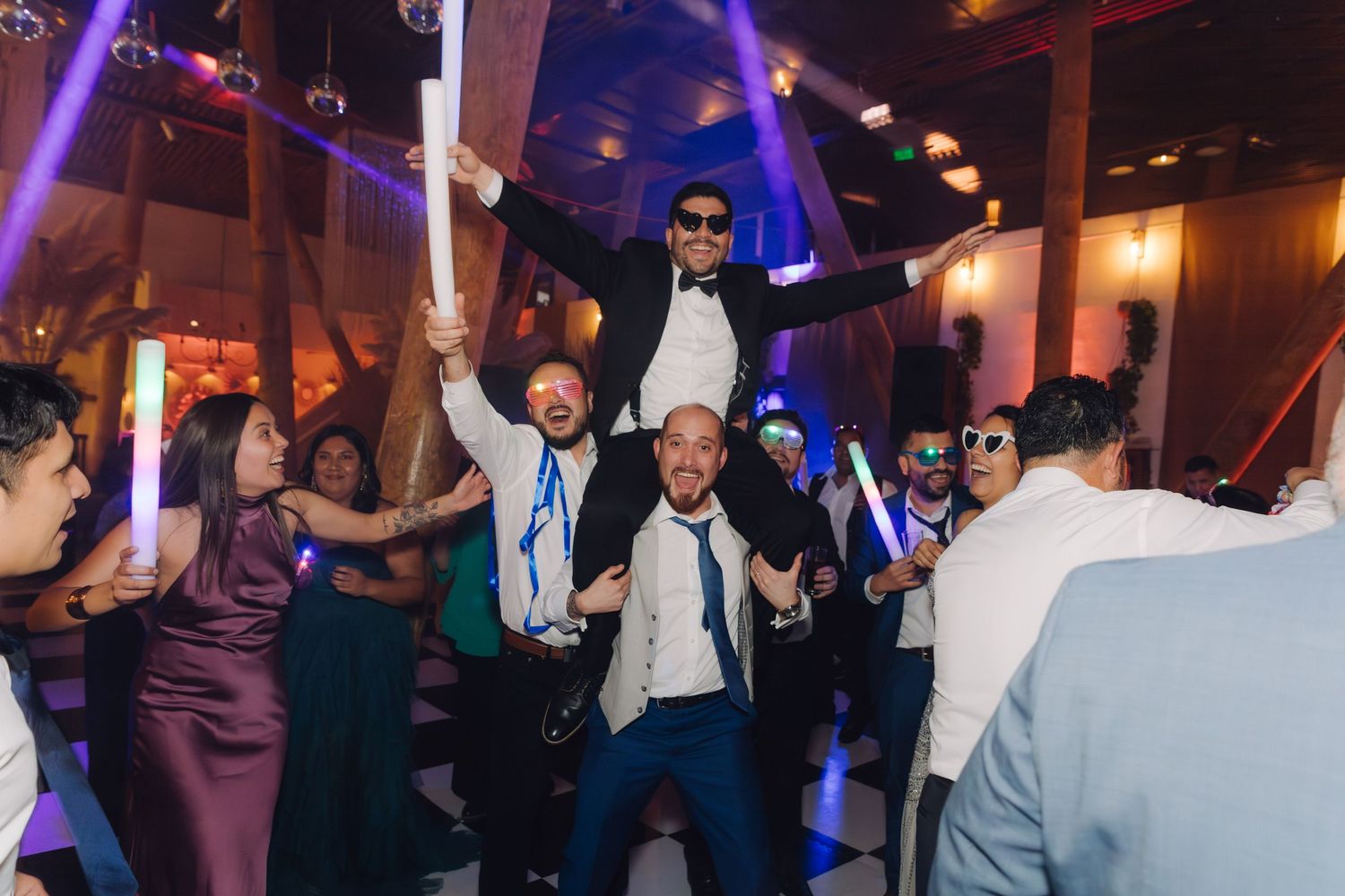 Groom lifted on shoulders by groomsmen during lively wedding reception dance party with colorful lights.
