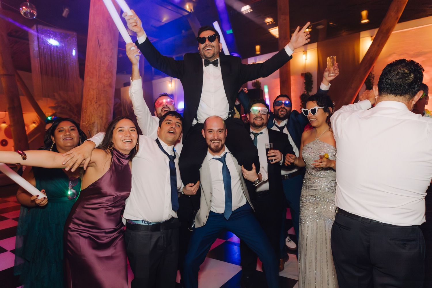 Wedding party group photo with groom elevated on shoulders celebrating on dance floor with purple lighting.