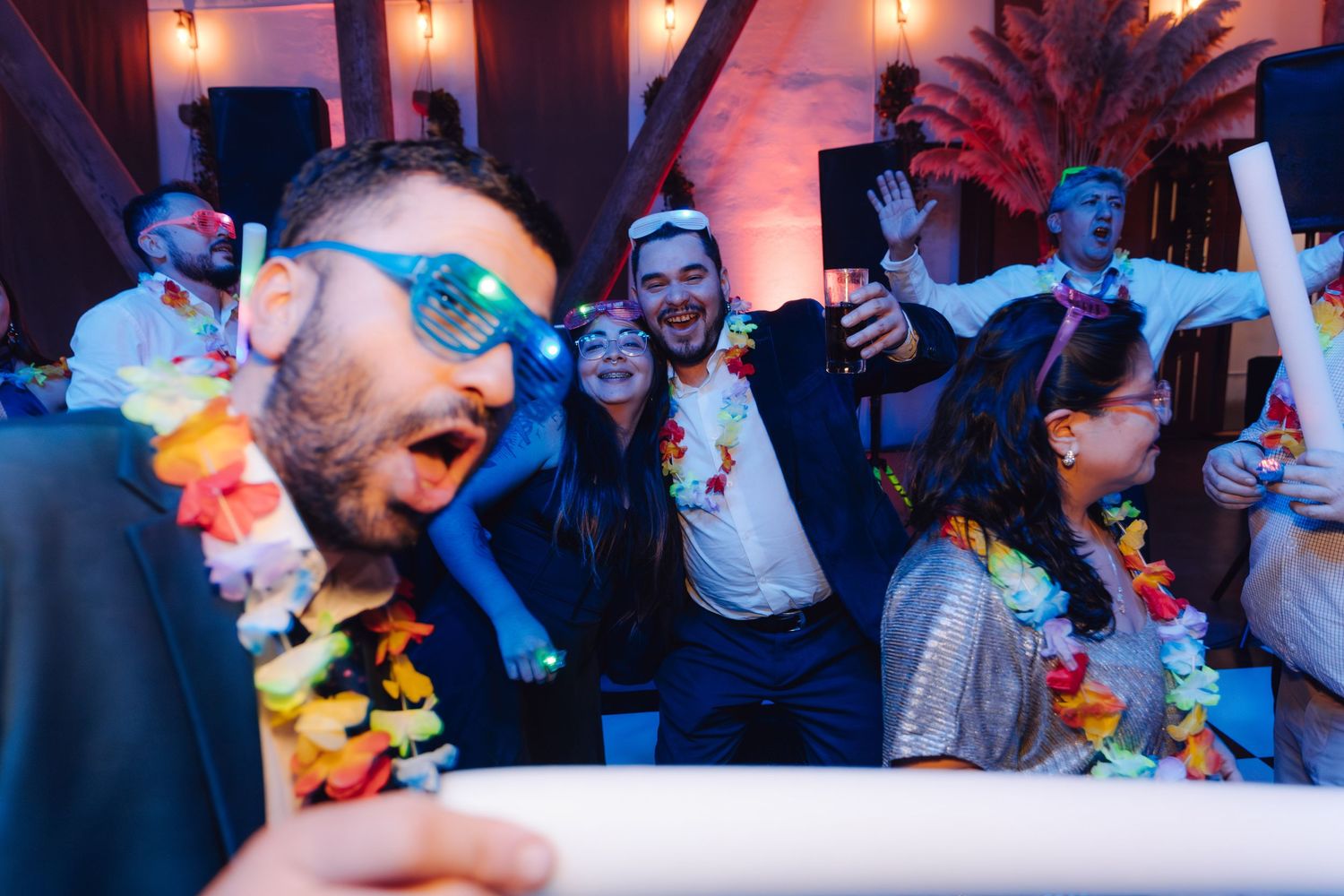 Wedding guests dancing and celebrating wearing Hawaiian leis and novelty glasses at festive reception party.