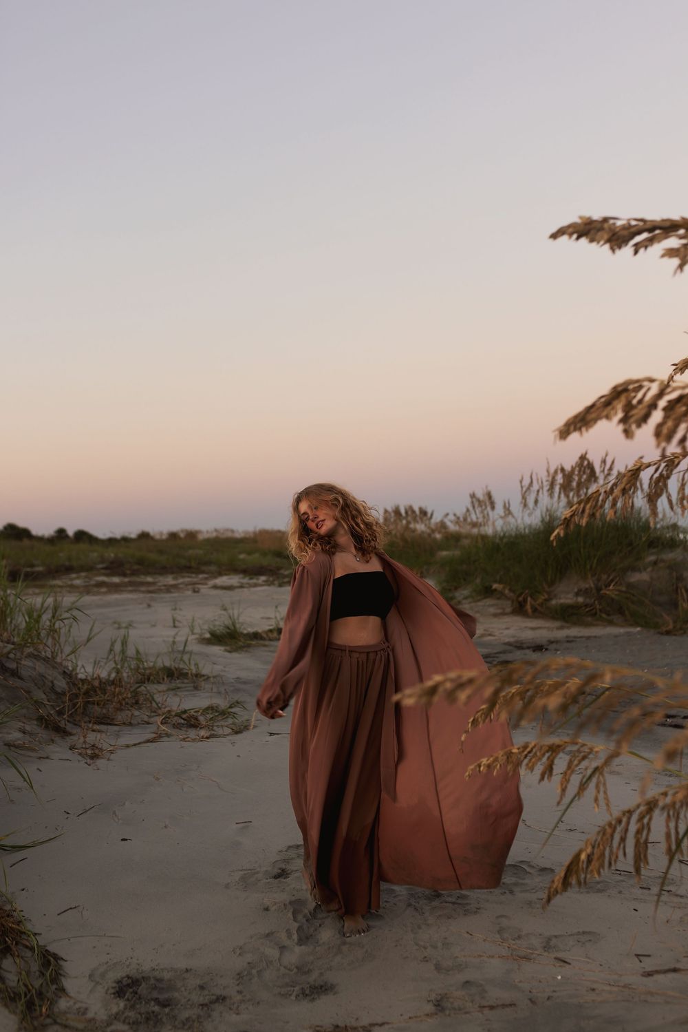 A series of beige beach chairs arranged along a sandy shore with sea oats and dunes at sunset.