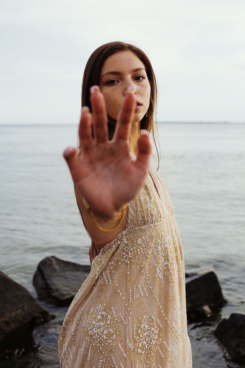 A person in a beige sequined dress stands near rocky shores with waves, raising their hand to block the camera view.