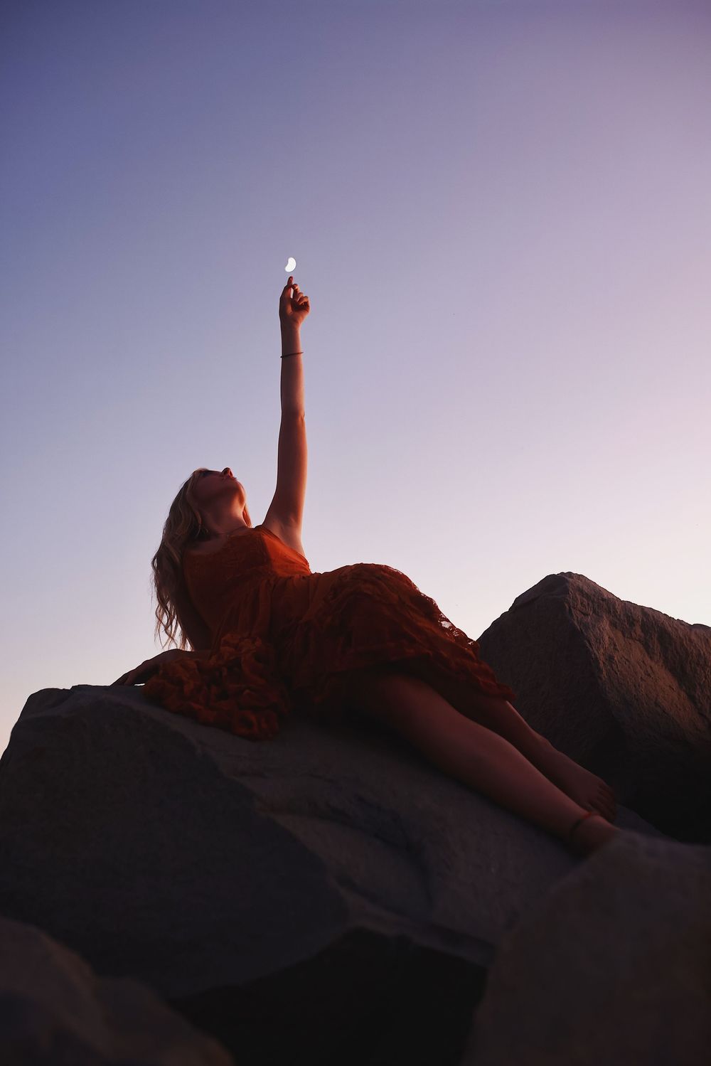 A silhouetted figure lies on rocks against a dusky purple sky with arm raised pointing upward.