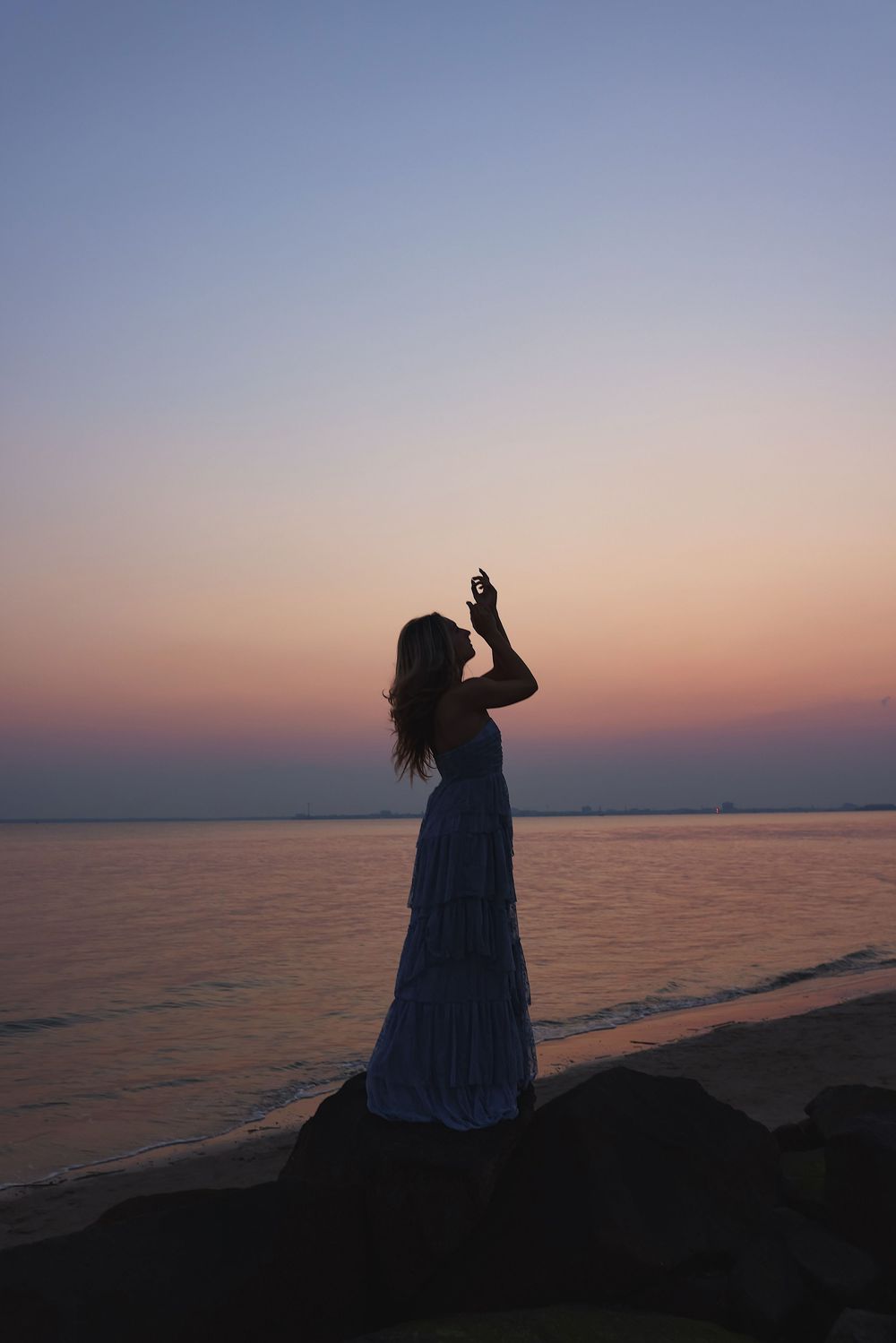Silhouetted figure sits on rocky beach watching colorful sunset over calm ocean waters.