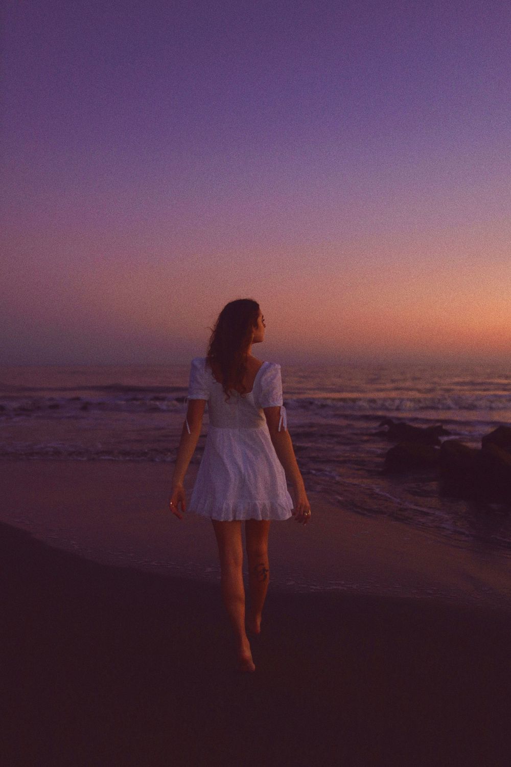 A person in a white dress walks along a beach at sunset creating a silhouette against the purple and orange sky.