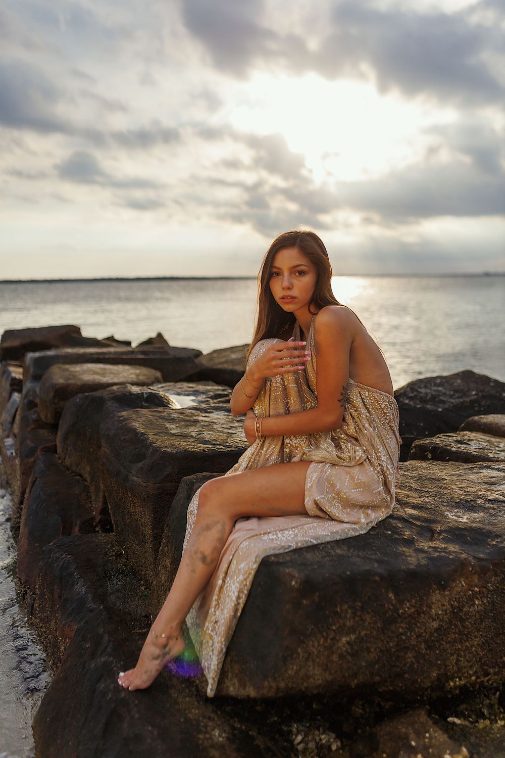 A person in a flowy dress poses on rocky coastal cliffs at sunset with dramatic cloudy skies over the ocean.