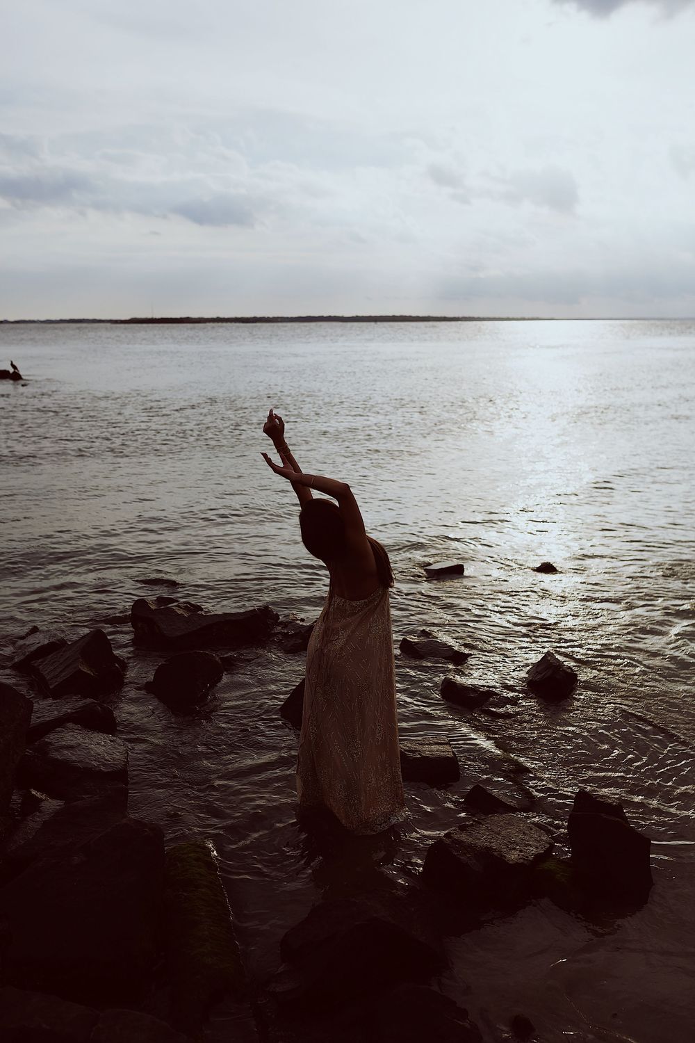 Silhouetted figure stretches gracefully against a moody seascape with rocky shoreline at dusk.