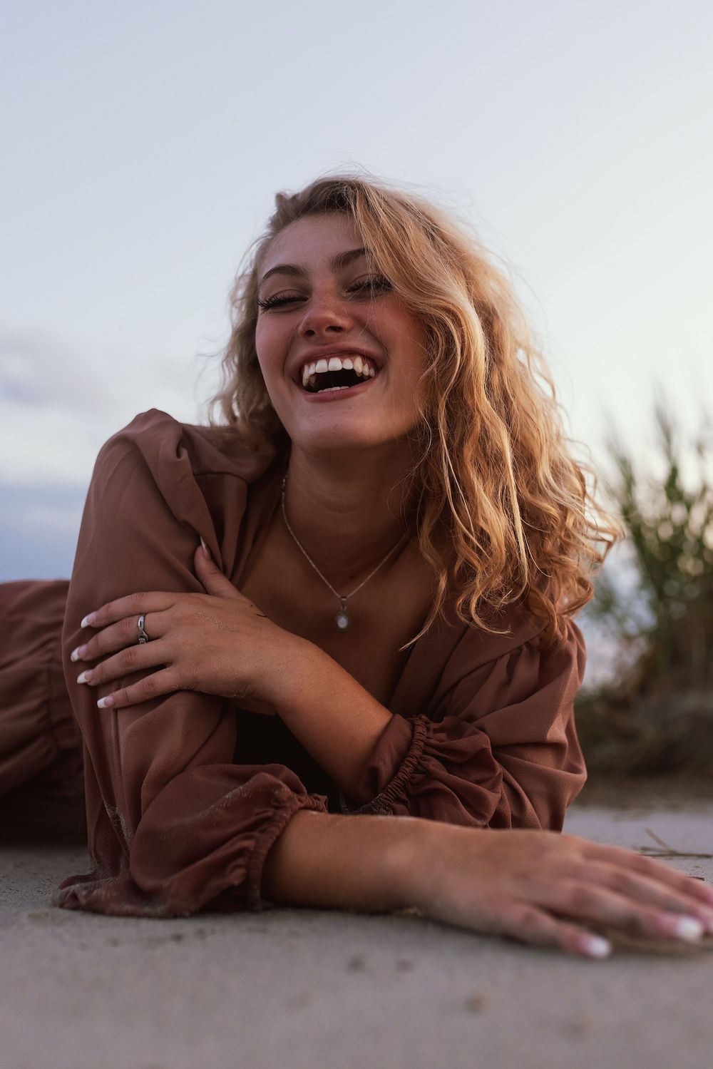 Joyful person with wavy blonde hair wearing brown dress lying down and laughing during golden hour photoshoot.