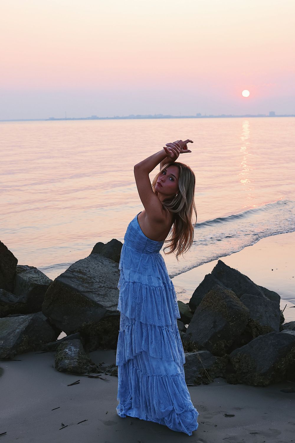 A person in a flowing blue dress poses dramatically against dark rocks at sunset by the ocean.