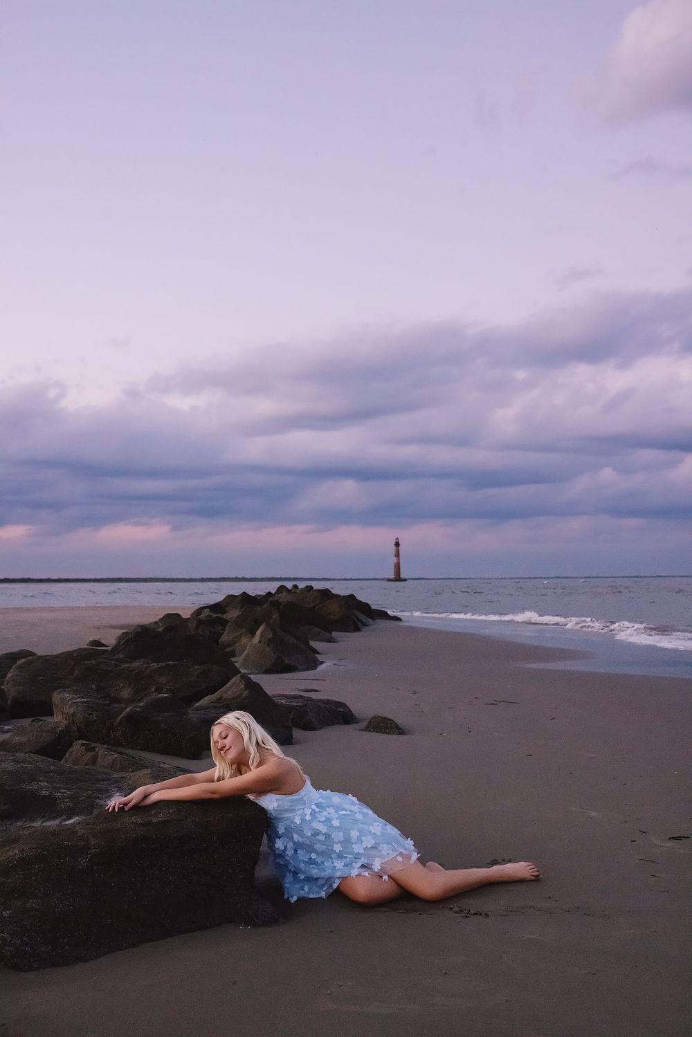A coastal scene at sunset with dramatic cloudy skies, rocky outcrops, and a sandy beach along the shoreline.